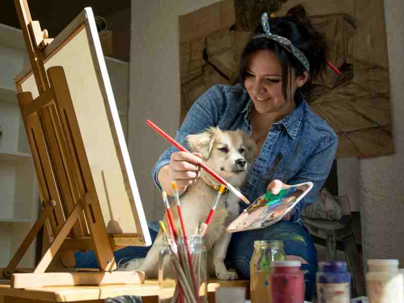 A woman paints a picture with her small dog at her side.